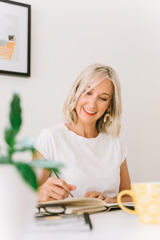 Copywriter (Australia) Kerrie Brooks smiling, sat at her desk writing in a notebook against a white background with a typewriter print on the wall. There is a yellow mug, plant and glasses in the foreground.