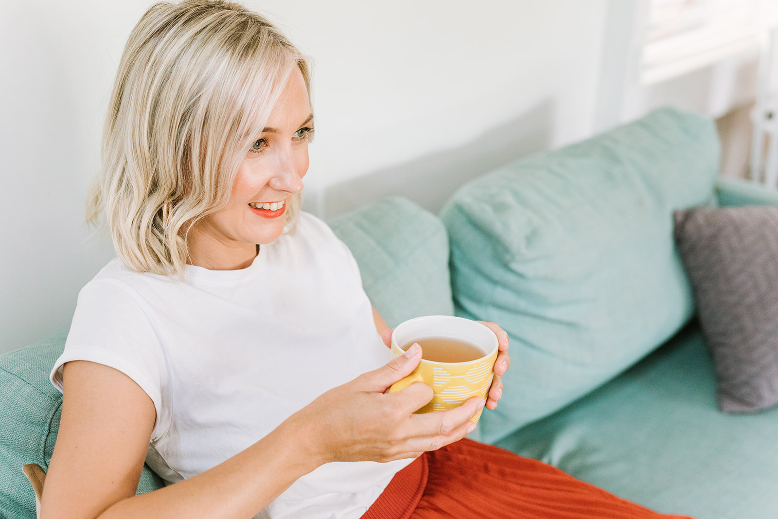 Newcastle Copywriter Kerrie Brooks wearing a white tee and rust-coloured skirt sat on a teal-coloured sofa holding a cup of tea in a yellow mug