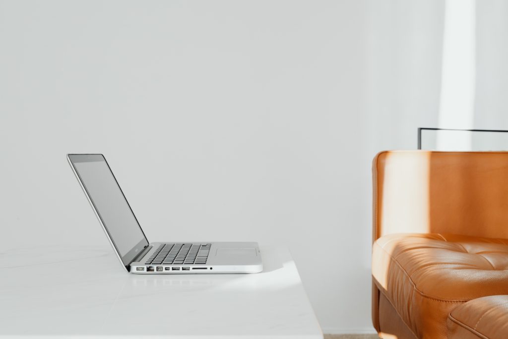 A sideways-facing open laptop sat on a white desk against a white wall. An orange retro leather lounge is in front of it.