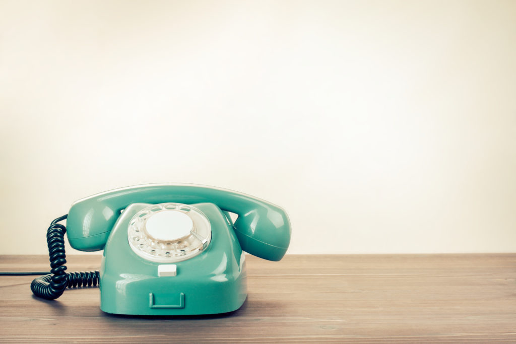 A teal-coloured retro dial phone sat on a wooden desk. Give KB a call!