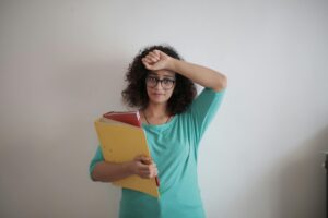 Woman with glasses in turquoise top holding folders in one hand and other hand on her brow 